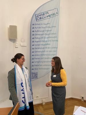Lea Schlenker und Mona Kuntze im Gespräch vor einer Fahne zur Charta Oecumenica