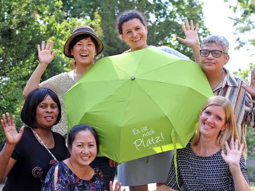 Das Fioto zeigt sechs Frauen unterschiedlicher Hautfarben mit einem aufgespannten Regenschirm auf dem steht Es ist noch Platz. Das Foto stammt von Es ist noch Platz, Foto Blutau, Weltgebetstag der Frauen Deutsches Komitee e.V.