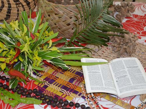 Zu sehen ist ein Foto mit Pflanzen aus den Gärten der Frauen in Vanuatu, eine Bislama-Bibel, Girlanden-Halsketten und palmengeflochtene Körbe und Fächer. 