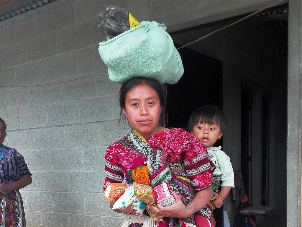 junge Frau mit Kind auf dem Rücken und Lebensmitteln unter den Armen und auf dem Kopf