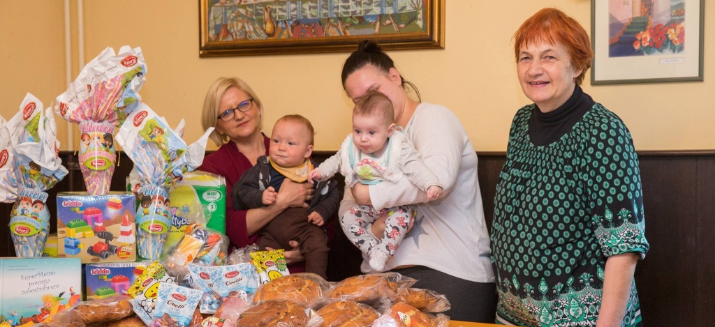 Zwei Frauen und ihre Babies sowie eine etwas ältere Frau stehen vor einem Tisch. Auf dem Tisch sind Spenden von Lebebsmitteln und Spielzeug von Spar Slowenien für das Mütterheim Pelikan der slowenischen Caritas, unterstützt vom Weltgebetstag. Das Foto stammt von Spar Slowenien 
